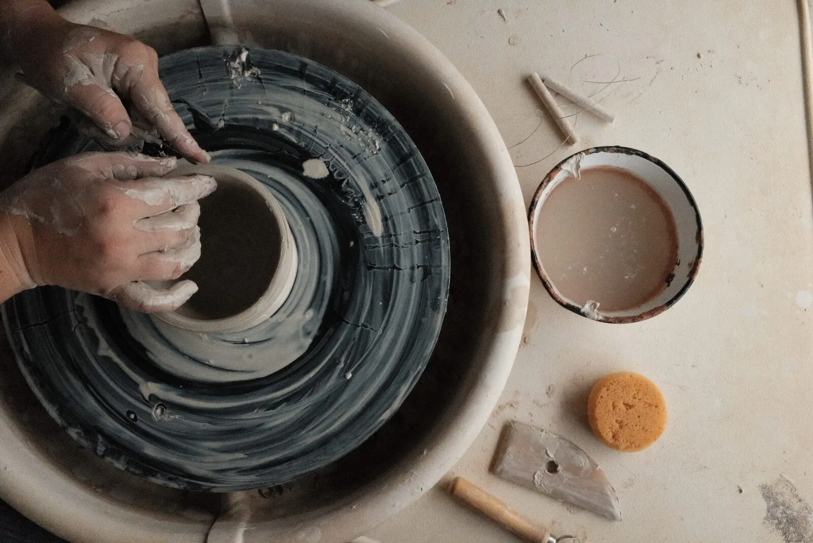 Clay-covered hands centering a pot on the wheel at a Devine Perks workshop, seen from above