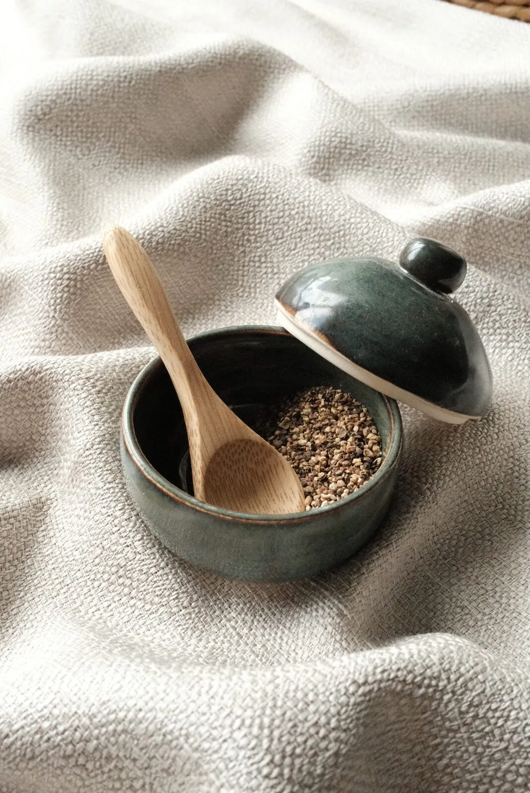 A hand-glazed lidded stoneware jar with a wooden spoon and grain, on soft linen