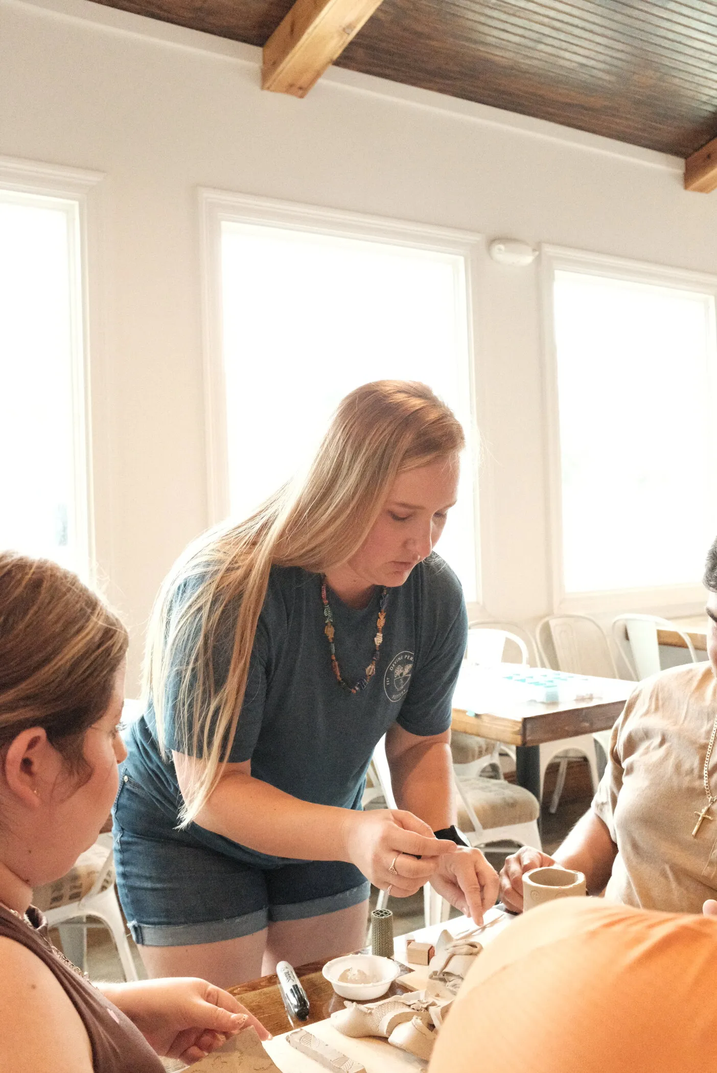Kelly Devine leading a hands-on pottery workshop, helping a student shape a piece at the table